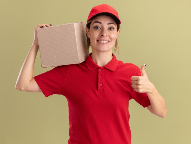 Sorridente giovane ragazza graziosa di consegna in uniforme pollice in alto e tiene cardbox sulla spalla su verde oliva