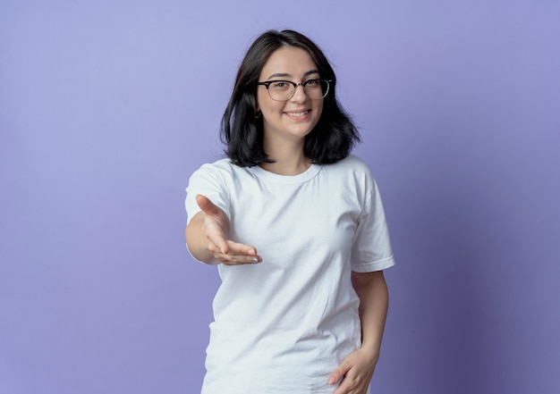 Sorridente giovane ragazza piuttosto caucasica con gli occhiali allungando la mano alla telecamera isolata su sfondo viola con spazio di copia