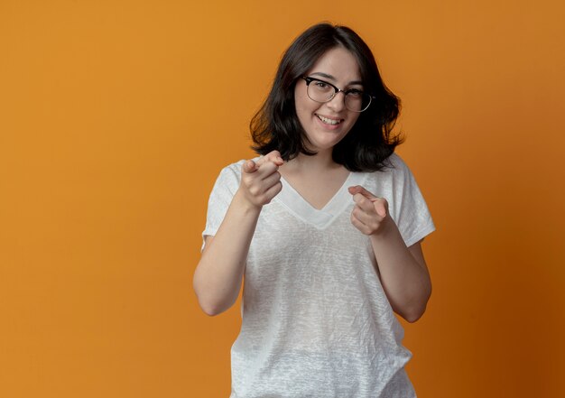 Sorridente giovane ragazza piuttosto caucasica con gli occhiali che punta alla telecamera isolata su sfondo arancione con copia spazio