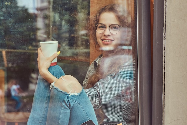 Sorridente giovane ragazza riccia rossa che indossa abiti casual e occhiali seduto su un davanzale con un caffè da asporto.