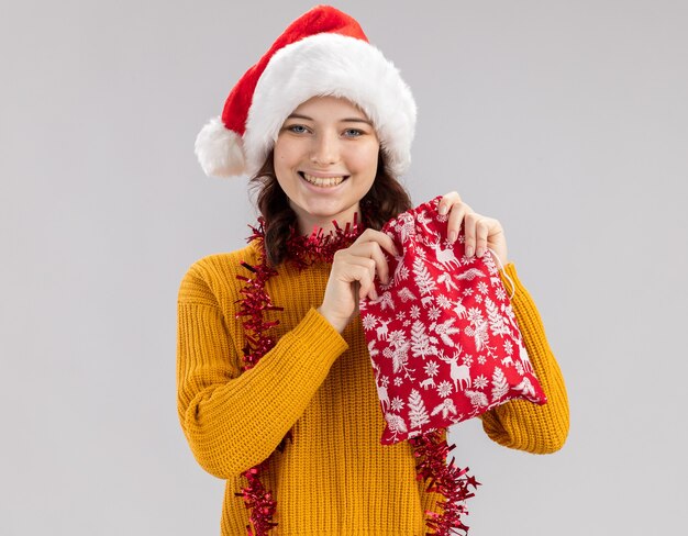 Sorridente giovane ragazza slava con cappello da Babbo Natale e con ghirlanda intorno al collo che tiene il sacchetto del regalo di Natale isolato sul muro bianco con copia spazio