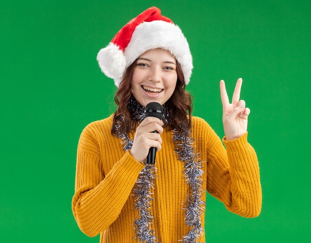 Sorridente giovane ragazza slava con cappello santa e con ghirlanda intorno al collo tenendo il microfono e gesticolando segno di vittoria