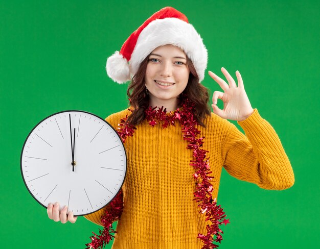 Sorridente giovane ragazza slava con santa hat e con una ghirlanda intorno al collo tenendo l'orologio e gesticolando segno ok isolato su sfondo verde con spazio di copia