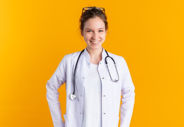 Sorridente giovane ragazza slava in uniforme medico con stetoscopio isolato sulla parete arancione con spazio copia