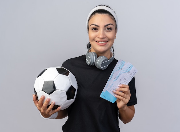Sorridente giovane ragazza sportiva caucasica che indossa fascia e braccialetti con le cuffie intorno al collo con biglietti aerei e pallone da calcio guardando davanti isolato sul muro bianco