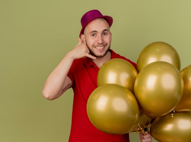 Sorridente giovane ragazzo bello slavo partito indossando il cappello del partito che tiene palloncini guardando la telecamera facendo gesto di chiamata isolato su sfondo verde oliva con lo spazio della copia