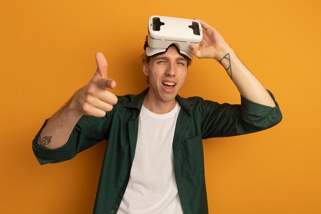 Sorridente giovane ragazzo biondo che indossa t-shirt verde e auricolare vr che ti mostra il gesto