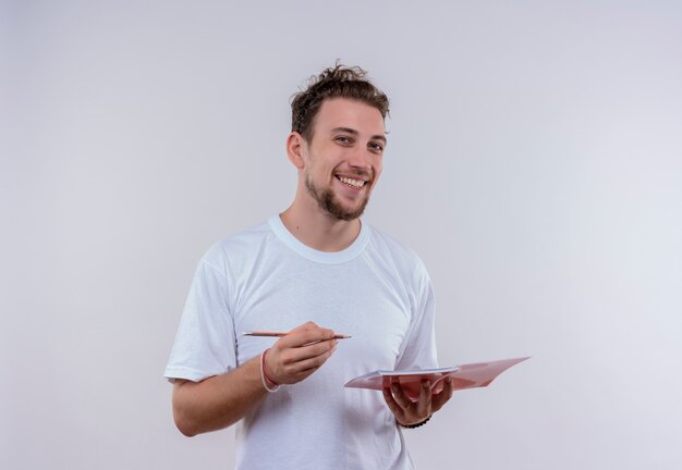 Sorridente giovane ragazzo che indossa t-shirt bianca tenendo matita e taccuino su sfondo bianco isolato