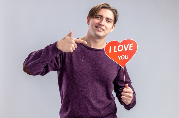 Sorridente giovane ragazzo il giorno di San Valentino che tiene e punta al cuore rosso su un bastone con ti amo testo isolato su sfondo bianco