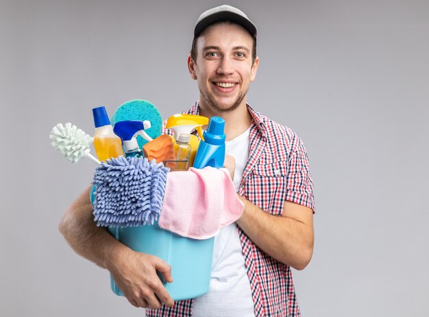 Sorridente giovane ragazzo pulitore che indossa cappello tenendo il secchio con strumenti di pulizia isolati su sfondo bianco white