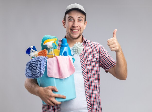 Sorridente giovane ragazzo pulitore che indossa il cappuccio tenendo la benna con strumenti di pulizia che mostra il pollice in alto isolato sul muro bianco