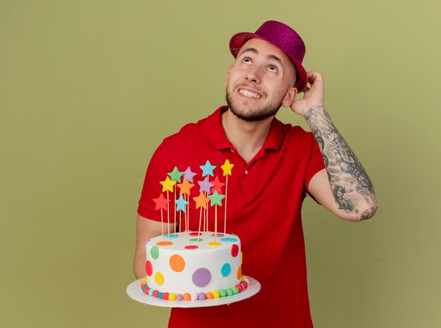 Sorridente giovane ragazzo slavo bello partito che indossa il cappello del partito che tocca la torta di compleanno della holding del cappello che osserva in su isolato su fondo verde oliva con lo spazio della copia