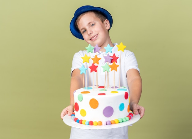 Sorridente giovane ragazzo slavo con blue party hat tenendo la torta di compleanno isolata sulla parete verde oliva con spazio copia