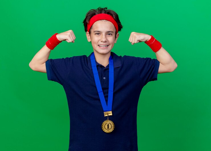 Foto gratuito sorridente giovane ragazzo sportivo bello che indossa fascia e braccialetti e medaglia intorno al collo con parentesi graffe guardando la parte anteriore facendo un forte gesto isolato sulla parete verde