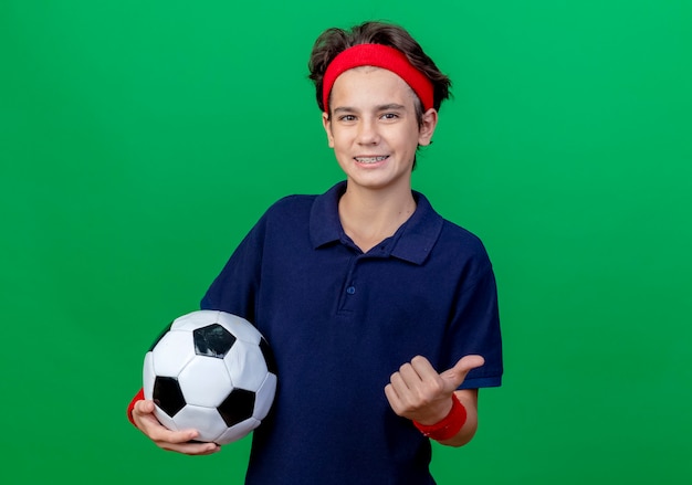 Sorridente giovane ragazzo sportivo bello che indossa la fascia e braccialetti con bretelle dentali che tiene il pallone da calcio che mostra pollice in su che guarda l'obbiettivo isolato su priorità bassa verde con lo spazio della copia