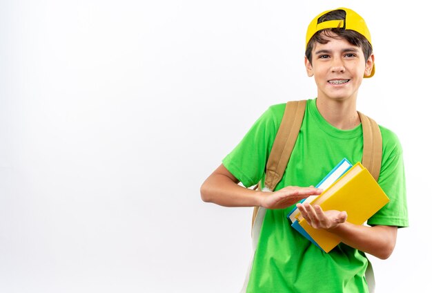 Sorridente giovane scolaro che indossa uno zaino con cappuccio che tiene libri isolati sul muro bianco con spazio per la copia