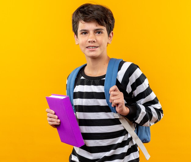 Sorridente giovane scuola ragazzo che indossa uno zaino tenendo il libro isolato sulla parete arancione