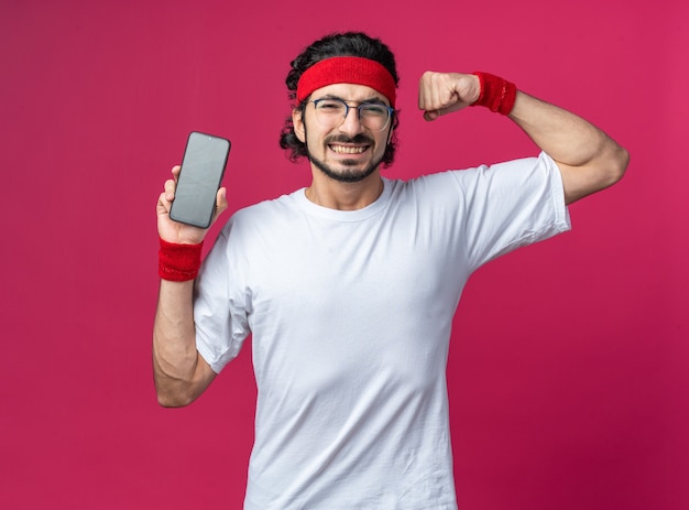 Sorridente giovane sportivo che indossa la fascia con il braccialetto che tiene il telefono che mostra un gesto forte