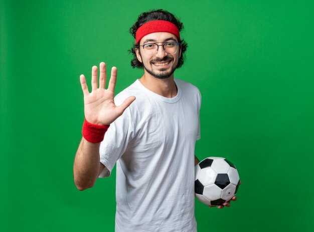 Sorridente giovane sportivo che indossa la fascia con il braccialetto che tiene la palla che mostra il gesto di arresto