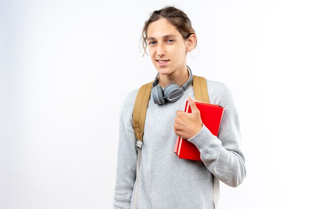 Sorridente giovane studente che indossa uno zaino con le cuffie sul collo che tiene libri isolati sul muro bianco con spazio di copia