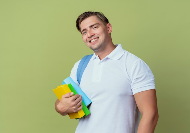 Sorridente giovane studente maschio bello che indossa la borsa posteriore che tiene i libri isolati su verde oliva