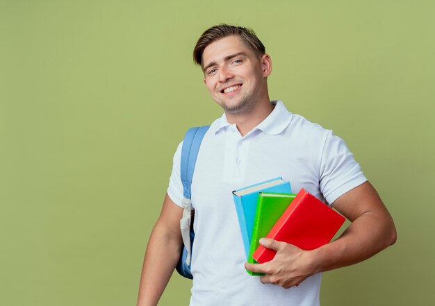 Sorridente giovane studente maschio bello che indossa la borsa posteriore che tiene i libri isolati su verde oliva