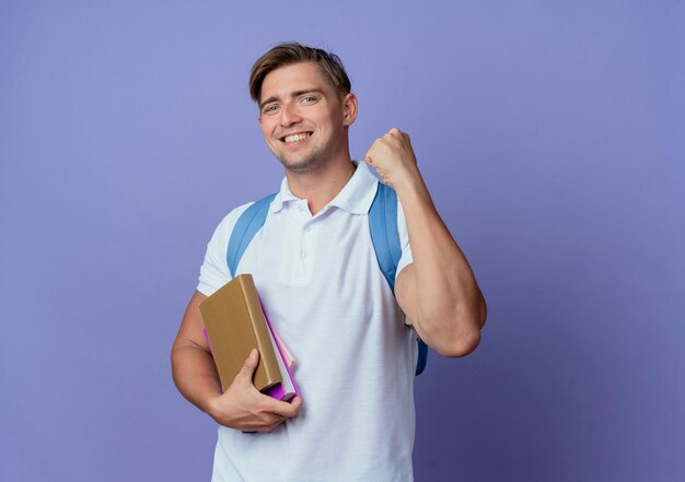 Sorridente giovane studente maschio bello che indossa la borsa posteriore in possesso di libri e mostrando sì gesto isolato sull'azzurro