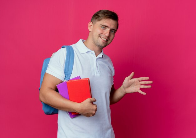 Sorridente giovane studente maschio bello che porta i libri della holding della borsa posteriore