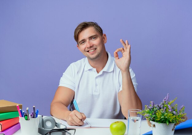 Sorridente giovane studente maschio bello seduto alla scrivania con strumenti di scuola che mostra il gesto okey e scrivere qualcosa sul taccuino isolato sull'azzurro