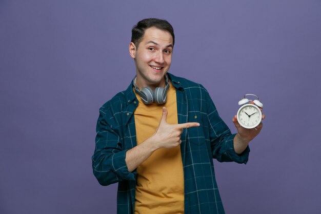Sorridente giovane studente maschio che indossa le cuffie intorno al collo che mostra la sveglia che punta verso di essa guardando la fotocamera isolata su sfondo viola