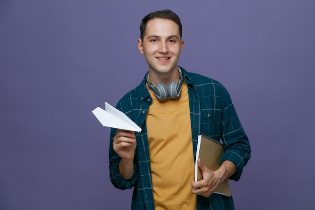 Sorridente giovane studente maschio che indossa le cuffie intorno al collo tenendo il blocco note sotto il braccio e aeroplano di carta guardando la fotocamera isolata su sfondo viola