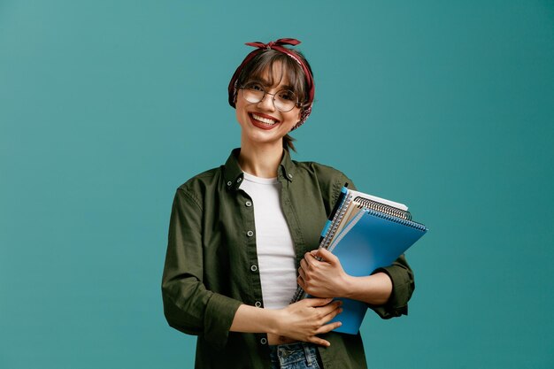 Sorridente giovane studentessa che indossa occhiali bandana tenendo grandi blocchi note con penna con entrambe le mani guardando la fotocamera isolata su sfondo blu