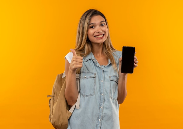 Sorridente giovane studentessa graziosa che indossa la borsa posteriore che mostra il telefono cellulare e il pollice in alto isolato sulla parete arancione