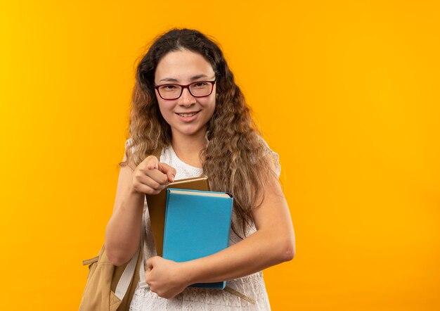 Sorridente giovane studentessa graziosa con gli occhiali e borsa posteriore che tengono i libri che indicano nella parte anteriore isolata sulla parete gialla