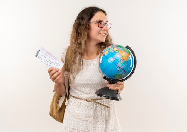 Sorridente giovane studentessa graziosa con gli occhiali e borsa posteriore che tiene globo e biglietti e guardando il lato isolato sul muro bianco