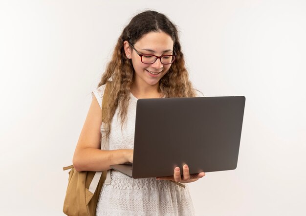 Sorridente giovane studentessa graziosa con gli occhiali e borsa posteriore utilizzando laptop isolato sulla parete
