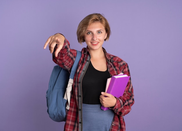 Sorridente giovane studentessa slava che indossa uno zaino con in mano un libro e un taccuino rivolto alla telecamera