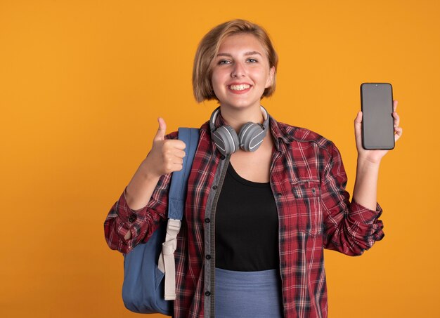 Sorridente giovane studentessa slava con le cuffie che indossa lo zaino tiene il telefono e il pollice in alto