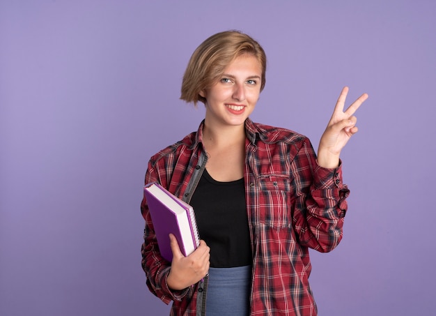 Sorridente giovane studentessa slava gesticola il segno della mano della vittoria tiene libro e taccuino