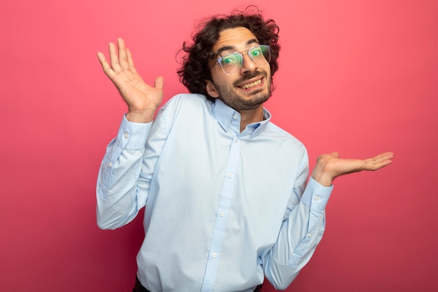 Sorridente giovane uomo bello con gli occhiali guardando la parte anteriore che mostra le mani vuote isolate sul muro rosa