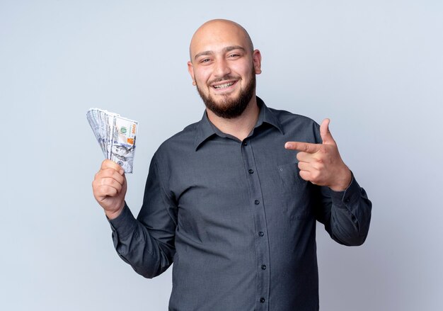 Sorridente giovane uomo calvo call center che tiene e che punta al denaro isolato sul muro bianco