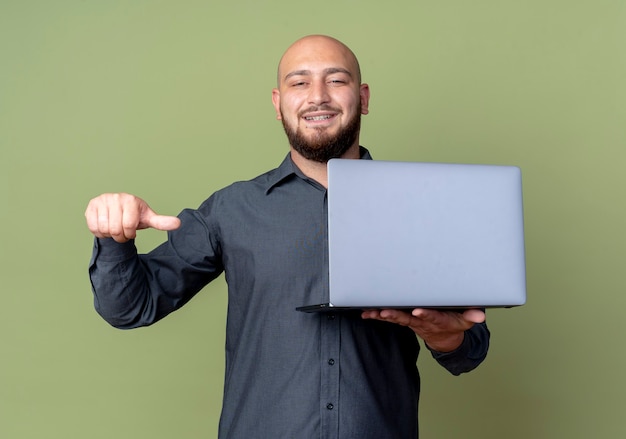 Sorridente giovane uomo calvo call center che tiene il computer portatile e indicandolo isolato sulla parete verde oliva
