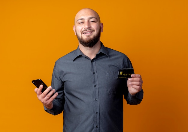Sorridente giovane uomo calvo call center in possesso di carta di credito e telefono cellulare isolato sulla parete arancione