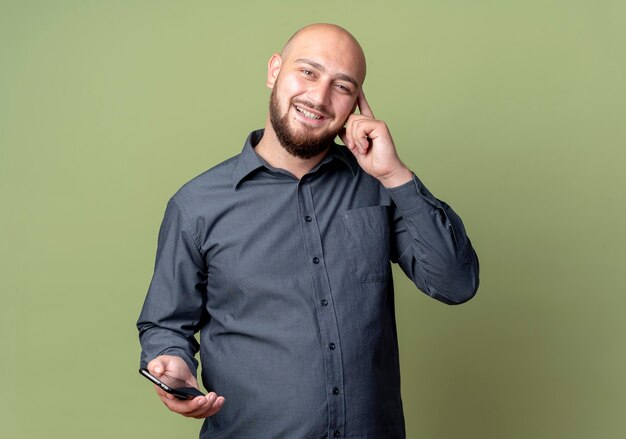 Sorridente giovane uomo calvo della call center che tiene il telefono cellulare che mette il dito sul tempio isolato sulla parete verde oliva