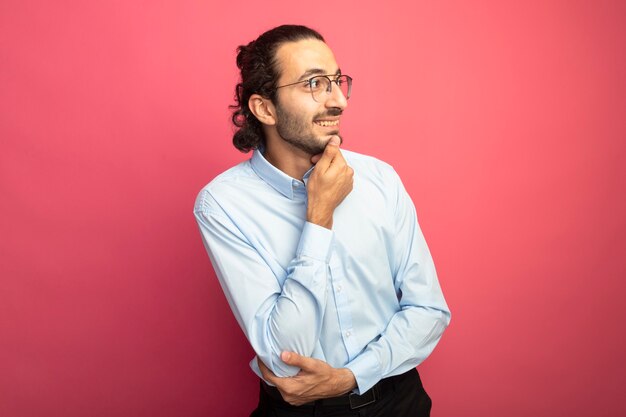 Sorridente giovane uomo caucasico bello con gli occhiali che tocca il mento guardando il lato isolato su sfondo cremisi con lo spazio della copia