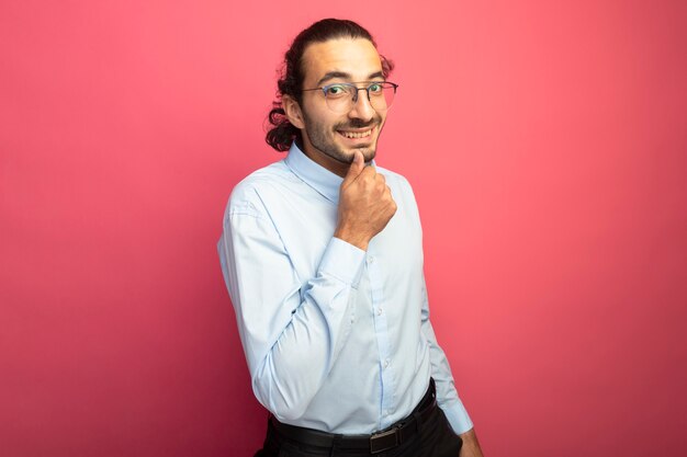 Sorridente giovane uomo caucasico bello con gli occhiali che tocca il mento guardando la telecamera isolata su sfondo cremisi con spazio di copia