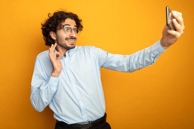 Sorridente giovane uomo caucasico bello con gli occhiali facendo segno ok tenendo selfie isolato su sfondo arancione