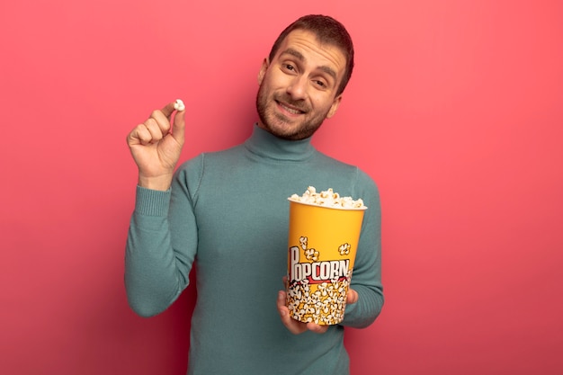 Sorridente giovane uomo caucasico guardando la telecamera tenendo il secchio di popcorn e popcorn pezzo isolato su sfondo cremisi con lo spazio della copia