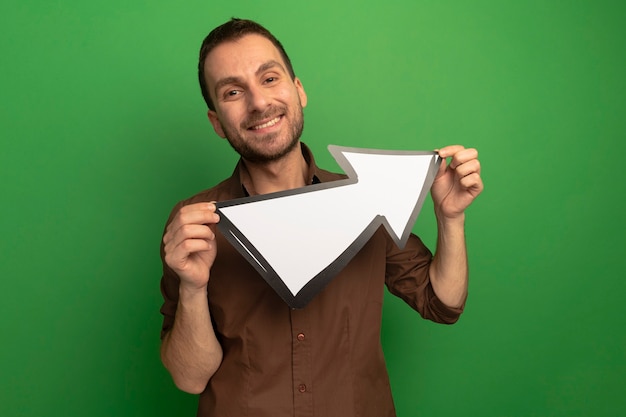 Sorridente giovane uomo caucasico guardando la telecamera tenendo premuto segno di freccia che punta a lato isolato su sfondo verde con spazio di copia