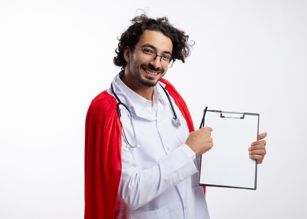 Sorridente giovane uomo caucasico supereroe in vetri ottici che indossa l'uniforme del medico con mantello rosso e con lo stetoscopio intorno ai punti del collo negli appunti tenendo la matita con lo spazio della copia